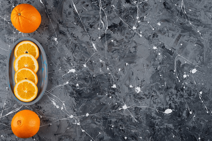 Overhead shot of a dark gray and white granite countertop, with two whole oranges and an oval dish of sliced oranges arranged on the left side.