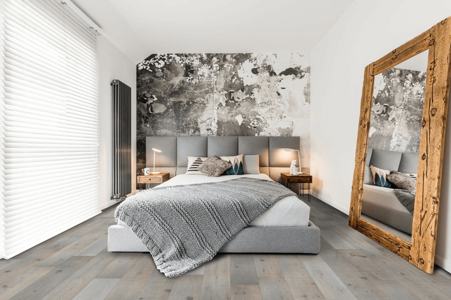 A modern bedroom featuring light gray wood-look flooring, a large bed with a gray headboard, an abstract accent wall, and a rustic full-length mirror.